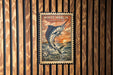 A vintage-style postage stamp featuring a large white marlin fish against a sunset background, mounted on a wooden wall.