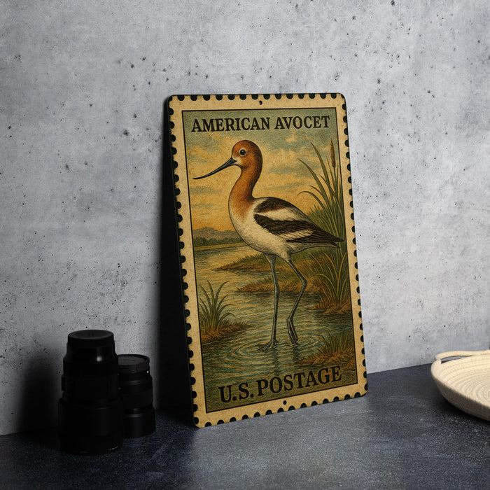 A metal sign with an illustration of a bird, likely a stork, standing in a body of water. The sign is framed with a border and has text that reads "AMERICAN AVOCET" and "U.S. POSTAGE".