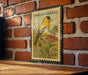 A vintage American Goldfinch postage stamp is displayed on a brick wall, featuring a yellow bird perched on a flower.