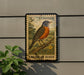 A postage stamp featuring an American robin is displayed on a wall, with a plant visible to the left.