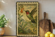 A framed postage stamp depicting a hummingbird is displayed on a shelf next to a wooden cutting board and some potted plants.