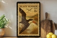 A framed postage stamp depicting a black skimmer bird is displayed on a shelf next to a wooden cutting board and a potted plant.