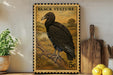 a framed stamp-like artwork depicting a black vulture perched on a branch, with a potted plant and a wooden cutting board visible in the background.