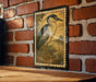 a framed illustration of a bird, likely a heron, mounted on a brick wall.