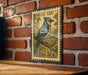 A vintage-style postage stamp featuring a blue jay perched on a branch, mounted on a brick wall.