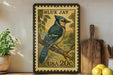 A framed postage stamp featuring a blue jay bird, with the text "BLUE JAY" and "USA 20¢" printed on it. The stamp is displayed on a shelf next to a wooden cutting board and some potted plants.
