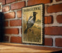 A vintage-style postage stamp featuring a black and white bird perched on a grassy field, mounted on a brick wall.