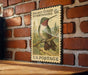 A vintage postage stamp featuring a colorful hummingbird perched on a branch, mounted on a brick wall.