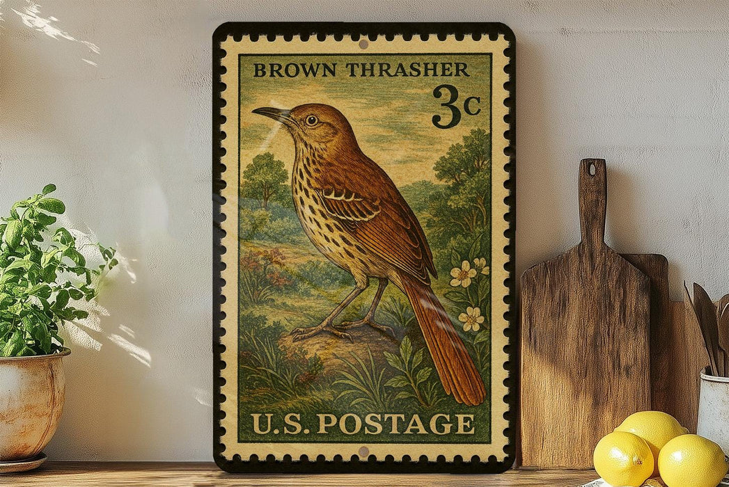A vintage brown thrasher postage stamp from the U.S. Postal Service, featuring a detailed illustration of a brown thrasher bird perched on a rock. The stamp is displayed on a shelf next to a wooden cutting board and a potted plant.