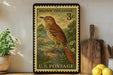 A vintage brown thrasher postage stamp from the U.S. Postal Service, featuring a detailed illustration of a brown thrasher bird perched on a rock. The stamp is displayed on a shelf next to a wooden cutting board and a potted plant.