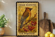 A vintage-style postage stamp featuring a bird perched on a branch with berries, displayed on a shelf next to a wooden cutting board and a potted plant.