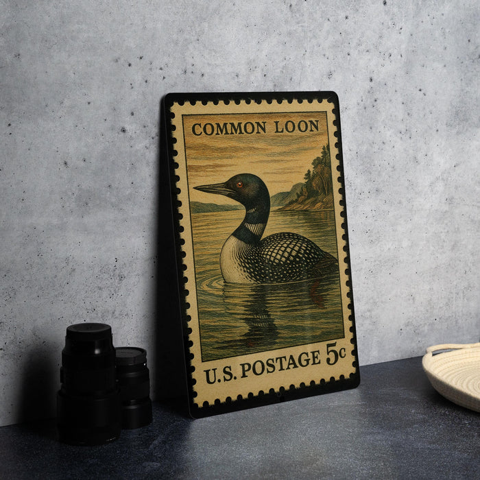 A framed postage stamp featuring a loon, a type of bird, is displayed on a table next to some black camera lenses.