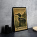 A framed postage stamp featuring a loon, a type of bird, is displayed on a table next to some black camera lenses.