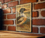 A vintage-style postage stamp featuring a duck is displayed on a brick wall.