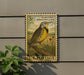 A U.S. postage stamp featuring a yellow bird perched on a branch, with the text "EASTERN MEADOWLARK" and "3c" printed on it. The stamp is mounted on a wall, and there is a plant visible to the left of the stamp.