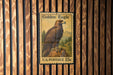 A U.S. postage stamp featuring a golden eagle perched on a rock, mounted on a wooden wall.