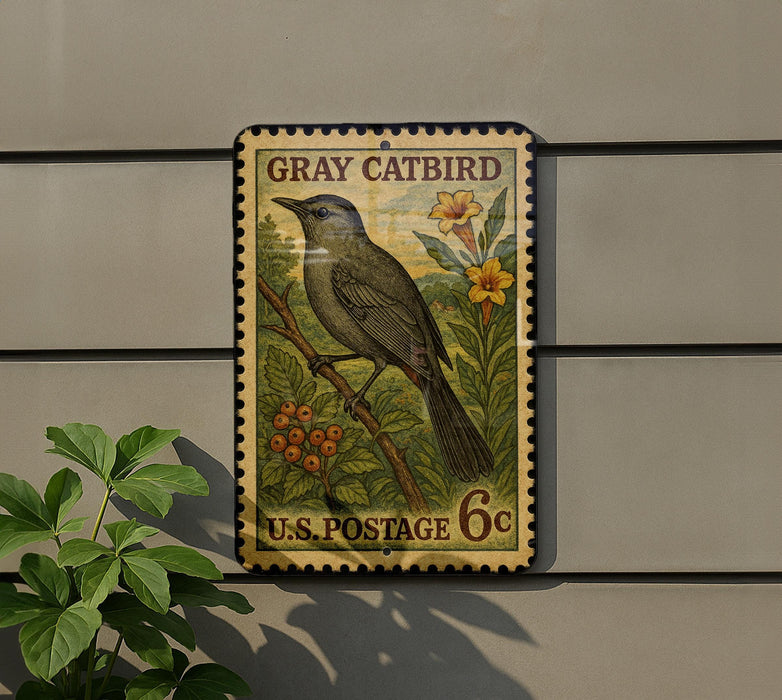 A vintage postage stamp featuring a gray catbird perched on a branch with flowers, mounted on a wall next to a plant.