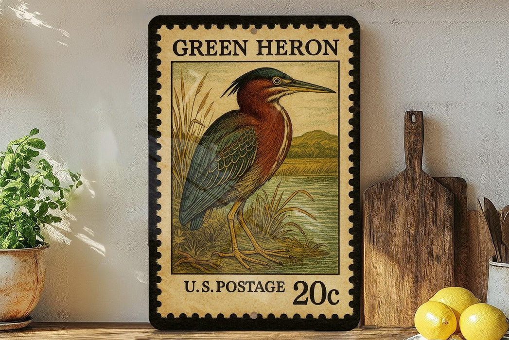 A framed postage stamp depicting a heron, with the text "GREEN HERON" and "U.S. POSTAGE 20c" printed on it. The stamp is placed on a shelf next to a wooden cutting board and a potted plant.
