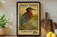 A framed postage stamp depicting a heron, with the text "GREEN HERON" and "U.S. POSTAGE 20c" printed on it. The stamp is placed on a shelf next to a wooden cutting board and a potted plant.