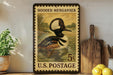 A framed postage stamp featuring a duck swimming in a pond, with the text "HOODED MERGANER 5¢ U.S. POSTAGE" printed on it.
