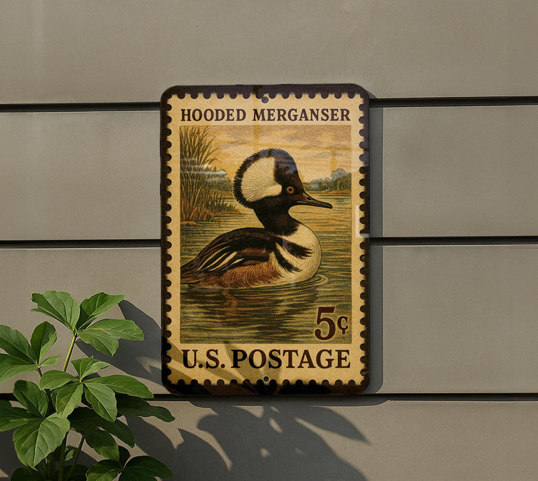 A vintage postage stamp featuring a hooded merganser duck, mounted on a wall next to a plant.