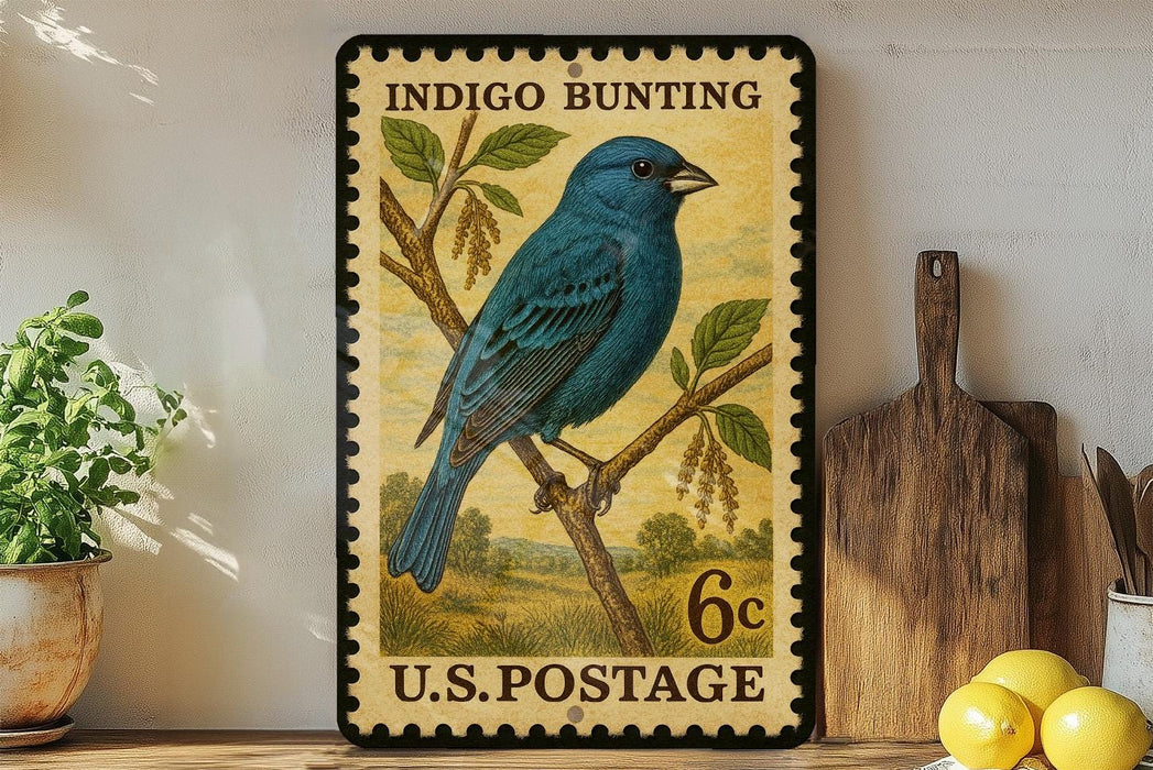 A vintage-style postage stamp featuring a blue bird perched on a branch, with the text "INDIGO BUNTING" and "U.S. POSTAGE" printed on it. The stamp is displayed on a shelf next to a wooden cutting board and a potted plant.