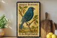 A vintage-style postage stamp featuring a blue bird perched on a branch, with the text "INDIGO BUNTING" and "U.S. POSTAGE" printed on it. The stamp is displayed on a shelf next to a wooden cutting board and a potted plant.