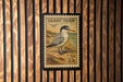 A framed postage stamp featuring a bird, likely a least tern, on a wooden background.