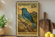 A framed U.S. postage stamp depicting a blue bird, likely a mountain bluebird, is displayed on a shelf next to a wooden cutting board and some lemons.