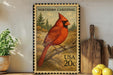 A framed postage stamp featuring a red cardinal perched on a branch, with a pine tree in the background. The stamp is from the United States and has a value of 20 cents.