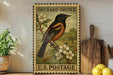 A vintage U.S. postage stamp featuring an orange oriole perched on a branch with white flowers, displayed on a shelf next to a wooden cutting board and a potted plant.