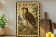 a framed stamp depicting an Osprey, a large bird of prey, perched on a branch. The stamp is placed on a shelf next to a wooden cutting board and a potted plant.