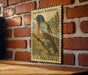 A vintage-style painted bunt bird postage stamp is displayed on a brick wall, with a wooden table below it.