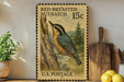 A framed U.S. postage stamp depicting a red-breasted nuthatch, placed on a wooden surface next to a wooden cutting board and a potted plant.
