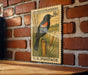 A vintage-style postage stamp depicting a blackbird perched on a reed plant, mounted on a brick wall.