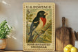 A U.S. postage stamp featuring a colorful illustration of a rose-breasted grosbeak perched on a branch, with a wooden post and a potted plant in the background.