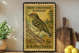 A vintage-style postage stamp featuring a yellow and green bird, likely a kinglet, perched on a branch. The stamp is displayed on a shelf next to a wooden cutting board and a potted plant.