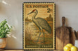 A vintage U.S. postage stamp featuring a sandhill crane is displayed on a shelf next to a wooden cutting board and a potted plant.