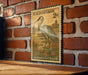 A vintage U.S. postage stamp featuring a sandhill crane is displayed on a wooden surface against a brick wall.