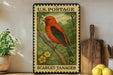 A vintage U.S. postage stamp featuring a red bird, likely a scarlet tanager, perched on a branch. The stamp is displayed on a shelf next to a wooden cutting board and a potted plant.