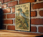 A framed postage stamp featuring a blue jay perched on a branch, mounted on a brick wall.