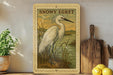 A framed illustration of a snowy egret standing in a grassy area, with a wooden cutting board and a potted plant to the side.