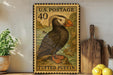 A framed postage stamp depicting a puffin bird, with the text "U.S. POSTAGE 40" and "TUFTED PUFFIN" printed on it. The stamp is displayed on a shelf next to a wooden cutting board and a potted plant.