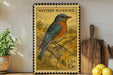 A framed postage stamp featuring a bluebird is displayed on a shelf next to a wooden cutting board and some potted plants.