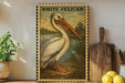 a framed illustration of a pelican, a large white bird with a long beak, standing on a grassy area near a body of water. The pelican is depicted in a detailed and realistic manner, with its distinctive features clearly visible. The framed illustration is placed on a shelf, surrounded by various kitchen utensils and items, such as a wooden cutting board, a knife, and a bowl.