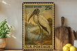 A vintage-style U.S. postage stamp featuring a wood stork, displayed on a shelf next to kitchen utensils and a potted plant.