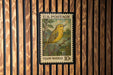 A U.S. postage stamp featuring a yellow warbler, a small songbird, is displayed on a wooden wall.