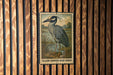 A metal stamp featuring a yellow-crowned night heron is displayed on a wooden wall.