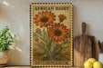 a vintage-style stamp or postcard with an illustration of two large orange daisy flowers, surrounded by greenery, placed on a wooden surface next to a wooden cutting board and a potted plant.