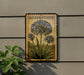 a framed illustration of blue flowers on a wall, with a plant to the left of the frame.